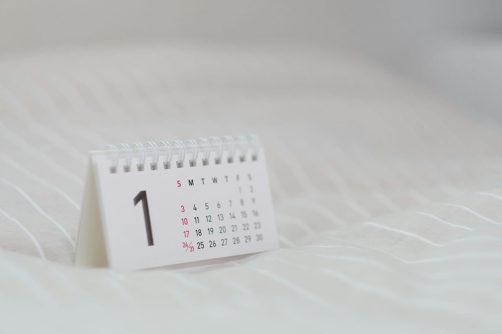 A small white spiral-bound desk calendar on striped fabric, with a large printed number 1 next to a month grid of dates 1 through 31
