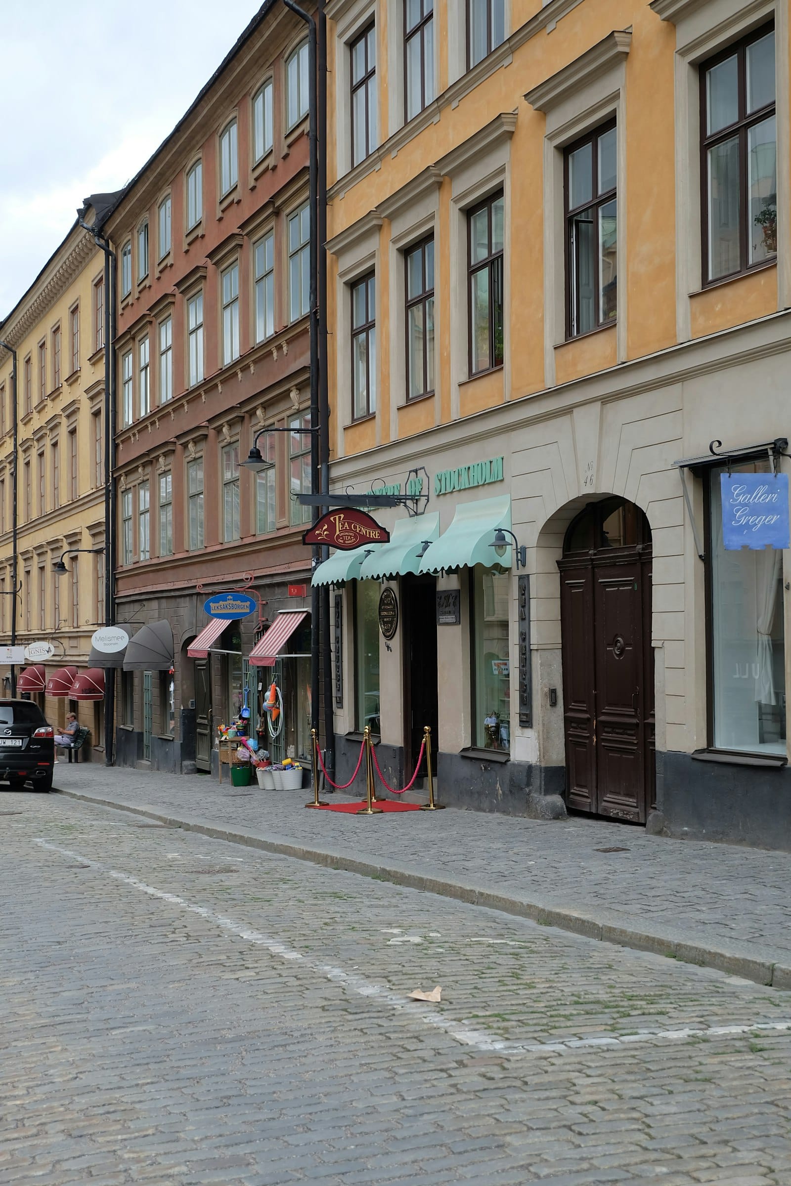 A cobblestone street of named shop fronts, tea shop, toy store, and gallery