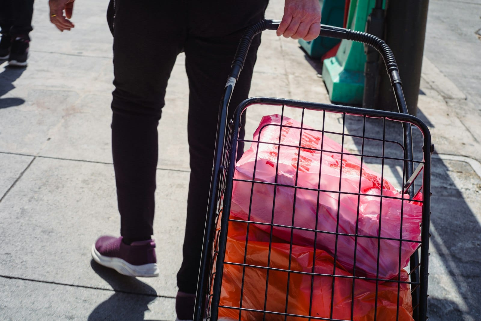 Person pulling a small shopping cart with bags on a sunny sidewalk