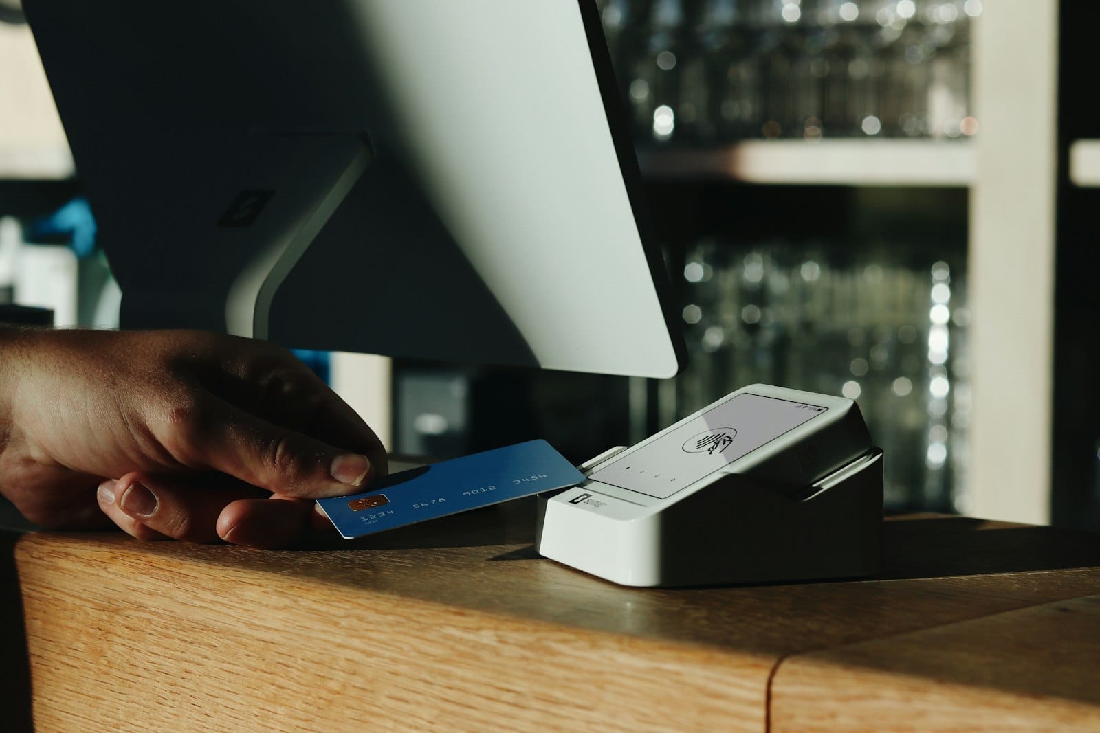 A hand holding a plain blue payment card at a card reader beside a computer screen