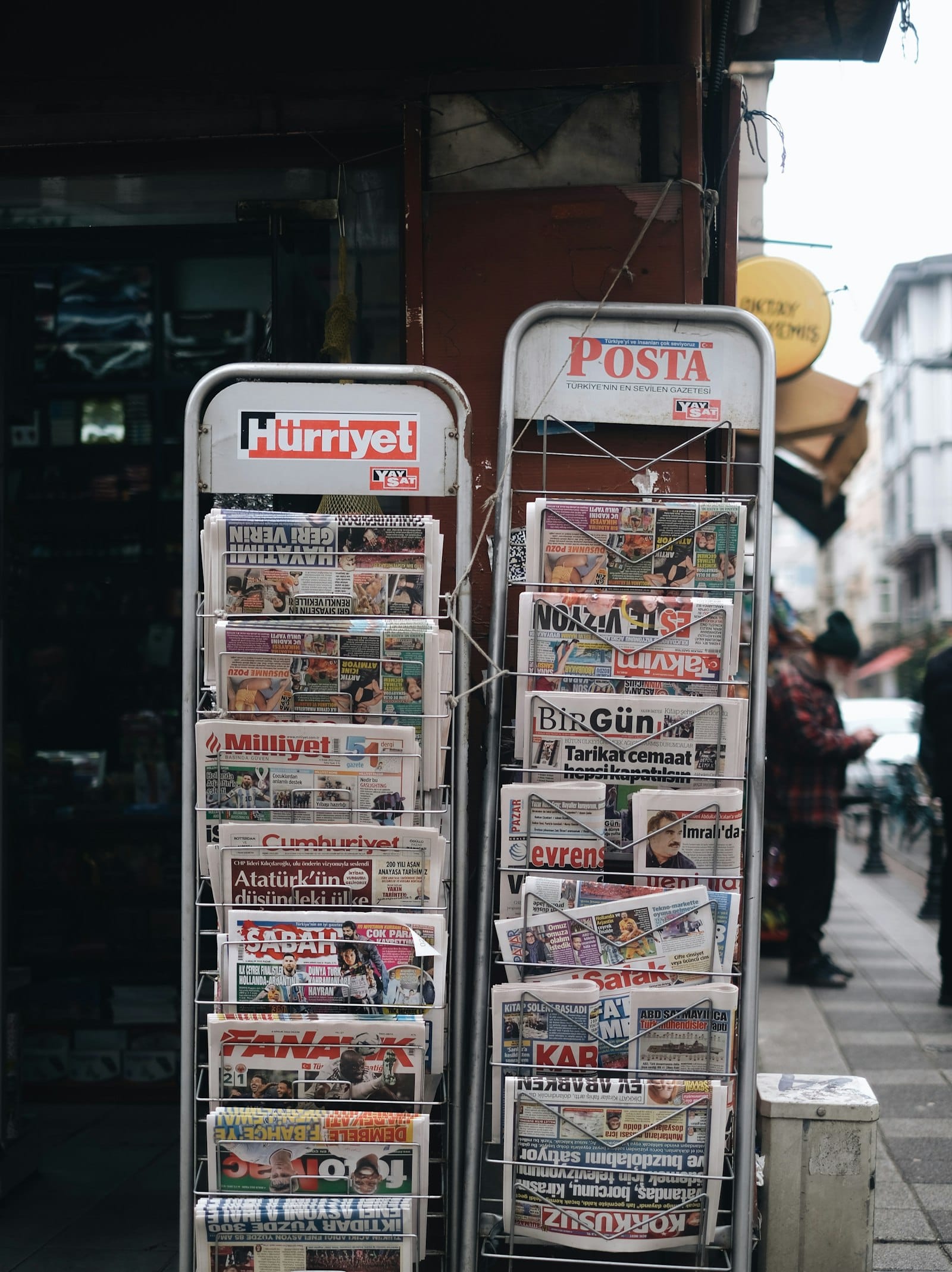 Two metal newspaper racks full of daily papers outside a corner shop, with a man walking past on the sidewalk