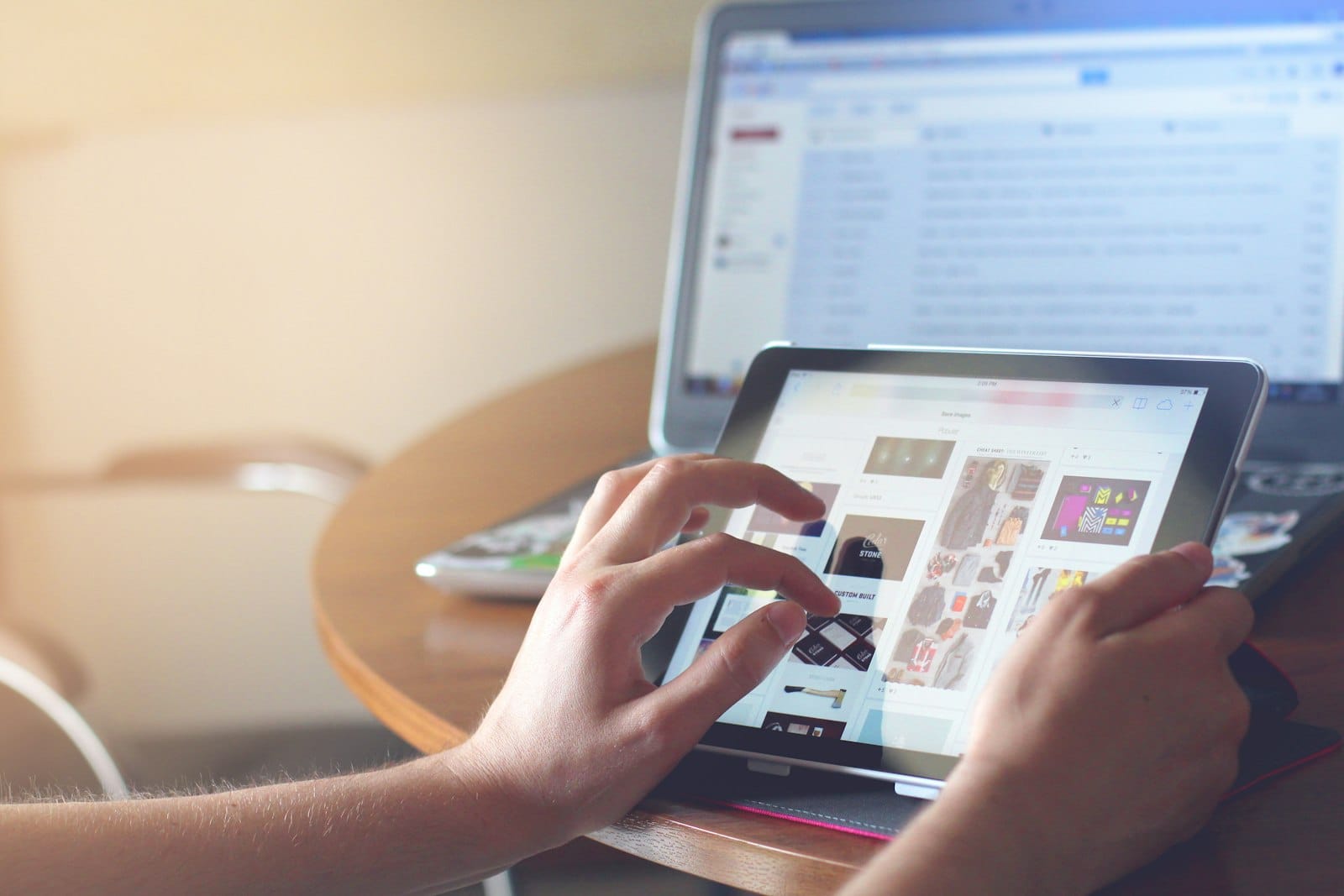 Hands swiping through a grid of photo thumbnails on a tablet, with a laptop open behind it