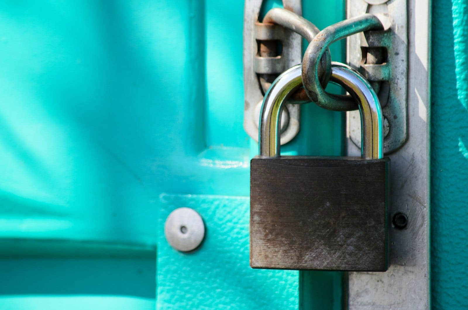 A dark padlock hanging from a latch on a turquoise door