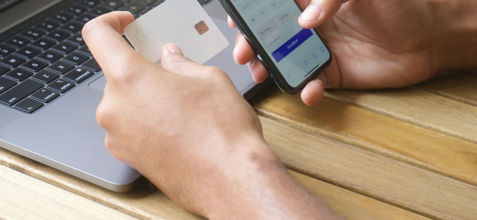 Hands holding a payment card beside a laptop keyboard while filling in a card form on a phone