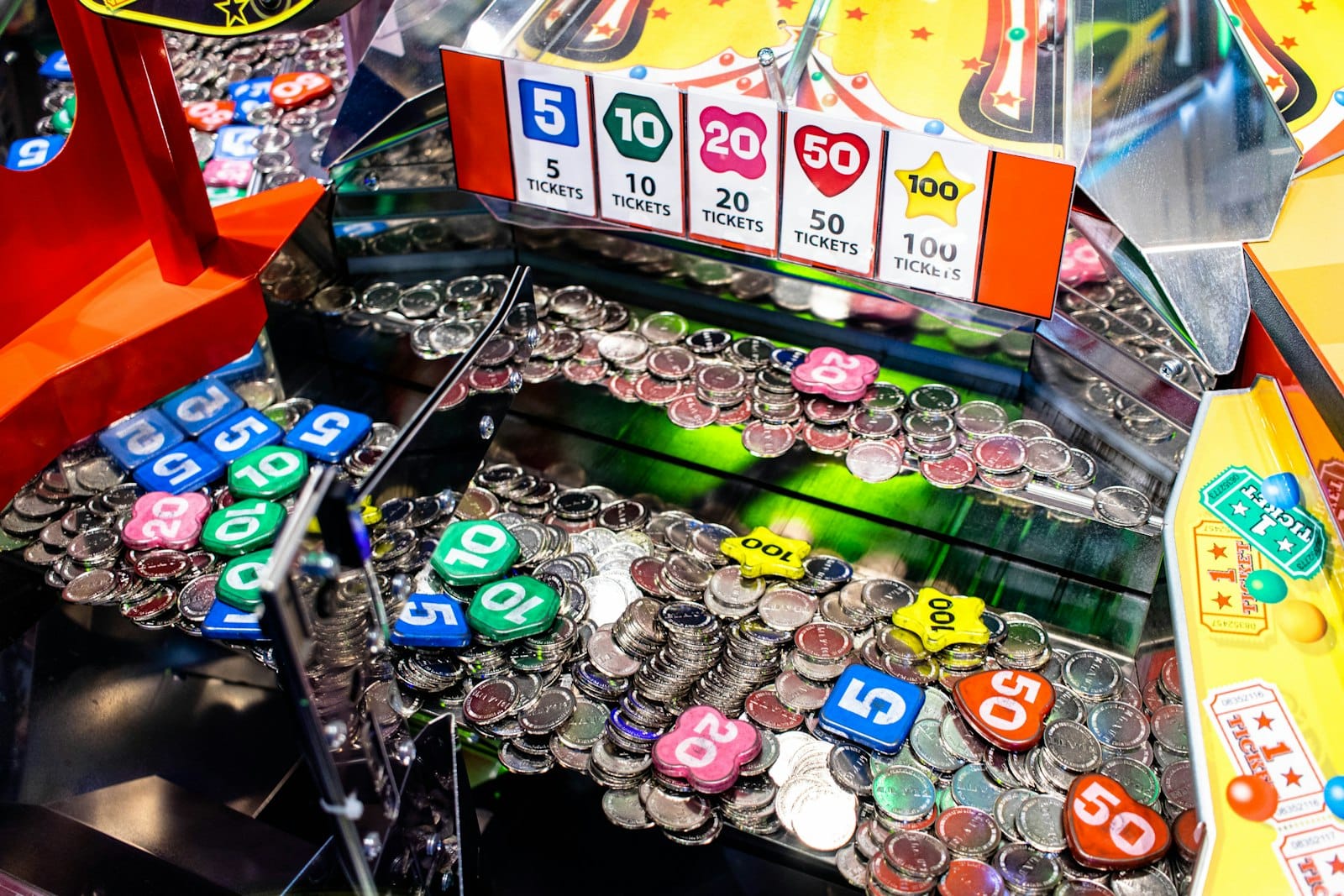 Coin pusher arcade machine full of silver coins and colorful numbered tokens worth 5 to 100 tickets